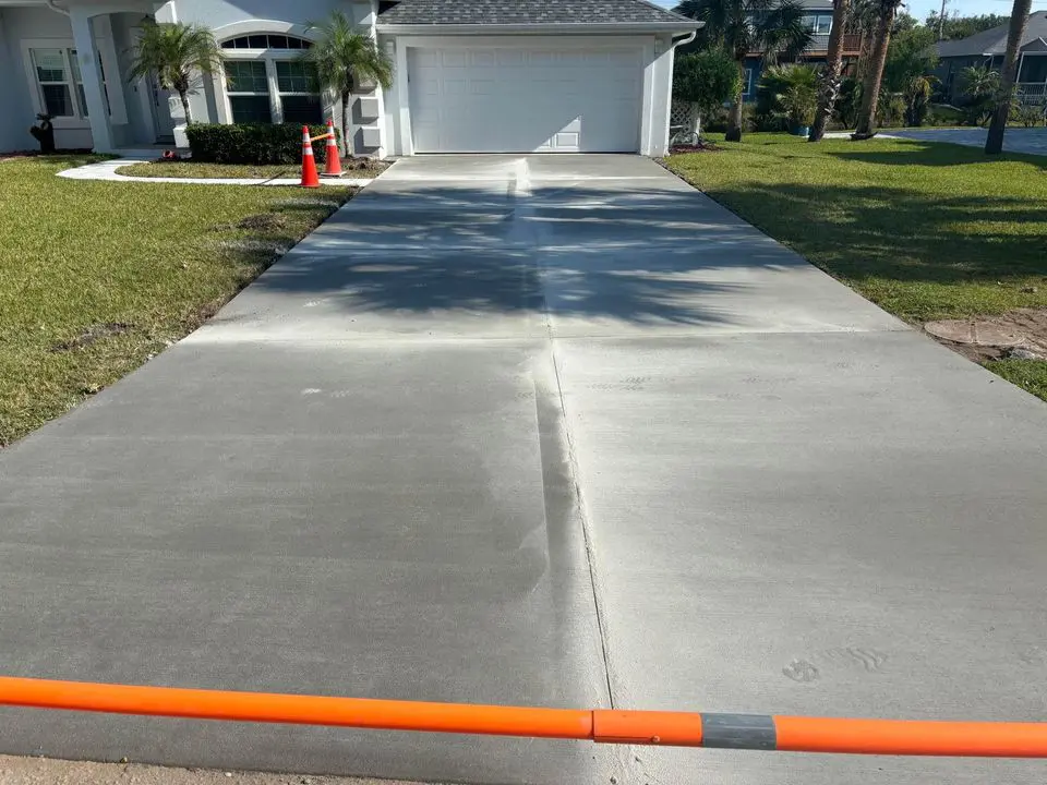 Concrete driveway which was poured and finished the day prior, has just had some saw cuts, also called expansion joints cut into the concrete driveway to stop it from cracking. The driveway is in front of a light blue house with a white garage door