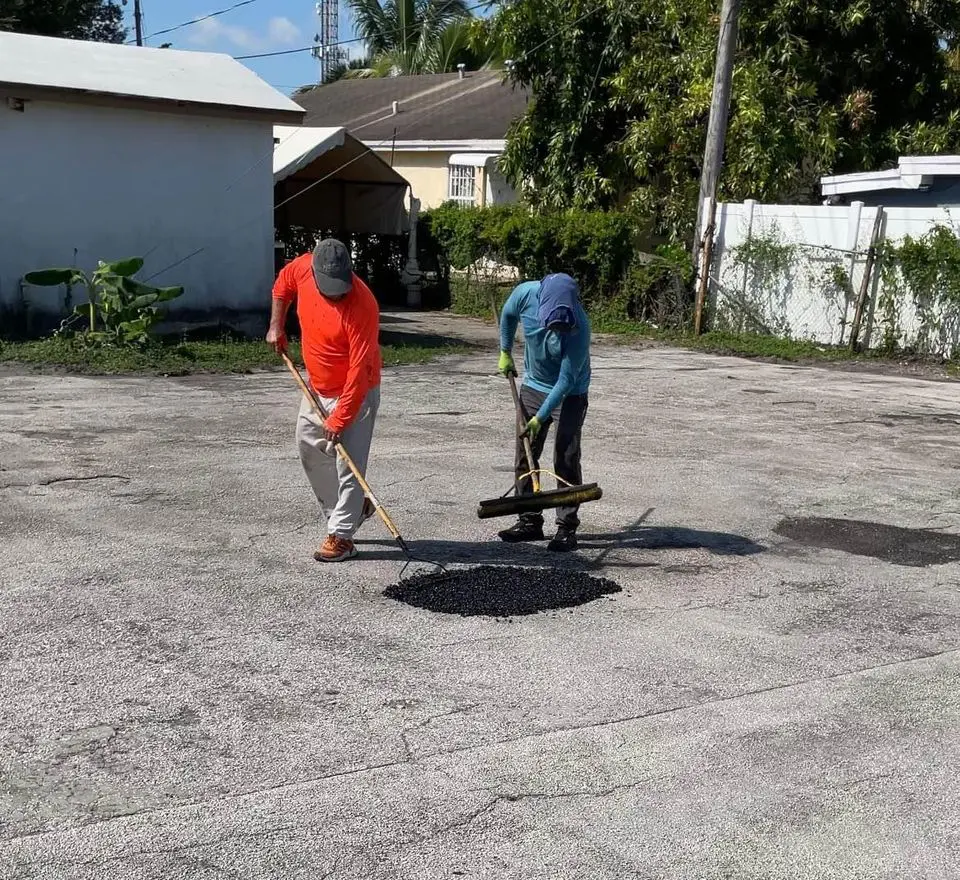 Two asphalt workers compacting a pothole in a small parking lot. Both asphalt contractors are looking down at the pothole they are working on.